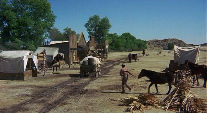 Still frame from a Western film. A horse-drawn wagon rides along a dirt road that stretches to the horizon. On one side of the road there are four small wooden buildings and two covered tents. The other side of the road is empty land.