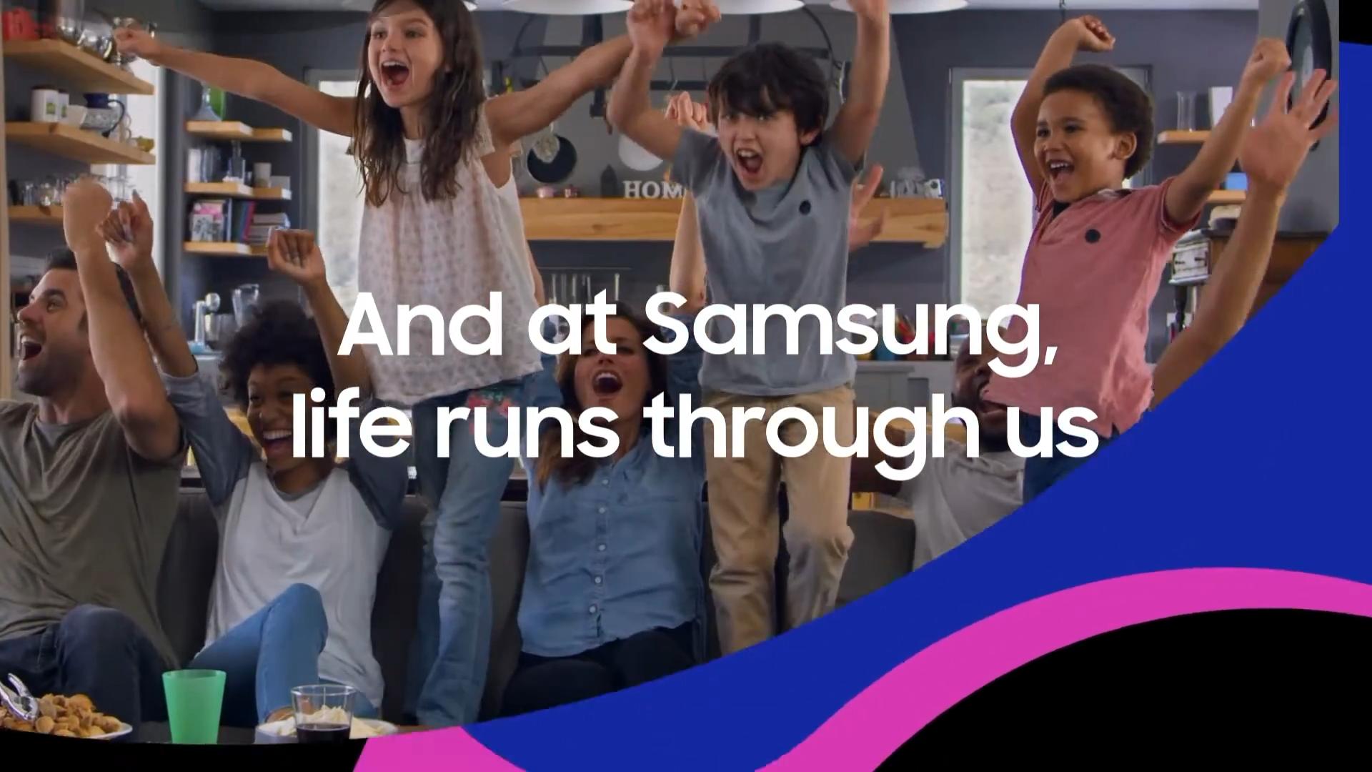 Still frame from Samsung's video. Four adults and three children are on the couch, excitedly smiling, jumping, and raising their hands in the air. Superimposed text reads "At Samsung, life runs through us".