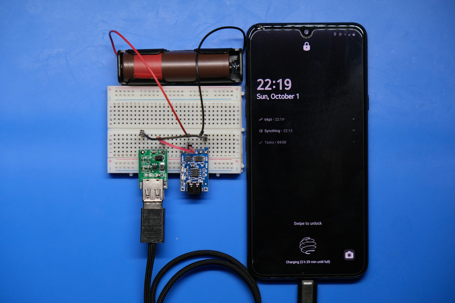Photograph of a breadboard containing a TP4056 charge controller, a 5 V USB regulator, and an 18650 battery cell. A cable connects this breadboard to a cell phone which is being charged.